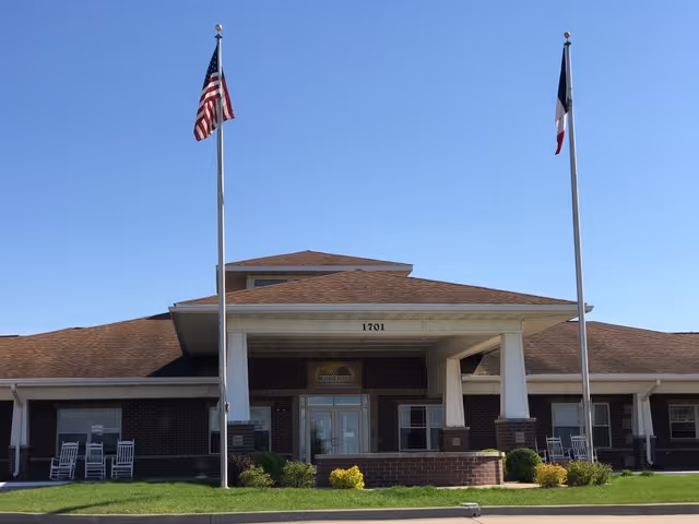 Front entrance of a brick senior living building with a covered portico, two flagpoles, and rocking chairs on the porch.