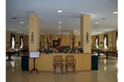 A spacious dining room with multiple tables and chairs arranged neatly. The room features beige walls with columns, wall-mounted light fixtures, and large windows with curtains allowing natural light to enter. There is a small stand with a menu or sign near the entrance area.