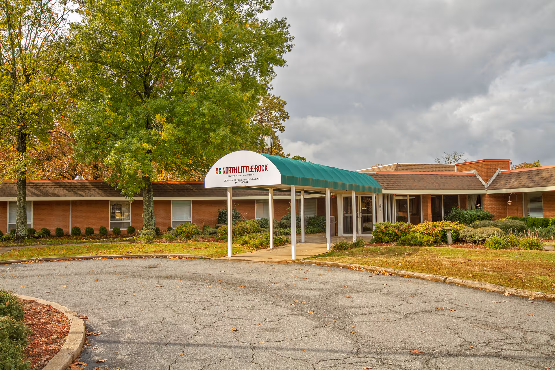 Exterior view of The Blossoms at North Little Rock Rehab & Nursing Center showing a single-story brick building with a green awning over the entrance. There are trees with green and autumn-colored leaves and a circular driveway in front of the entrance.