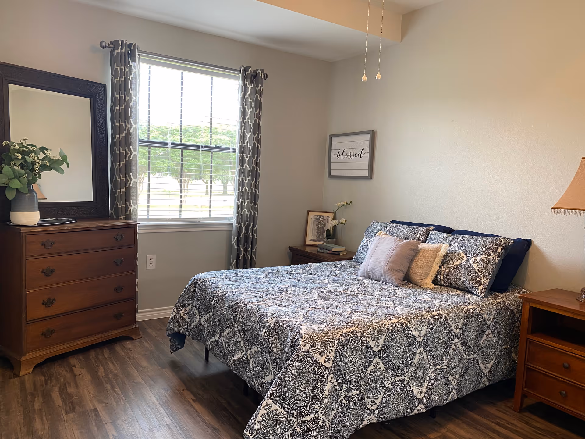 A cozy bedroom with a large window covered by patterned curtains. The bed has a blue and white patterned quilt with multiple pillows. There is a wooden dresser with a large mirror and a vase with greenery on top. A nightstand with a lamp and framed pictures is beside the bed. The room has wooden flooring and light-colored walls.