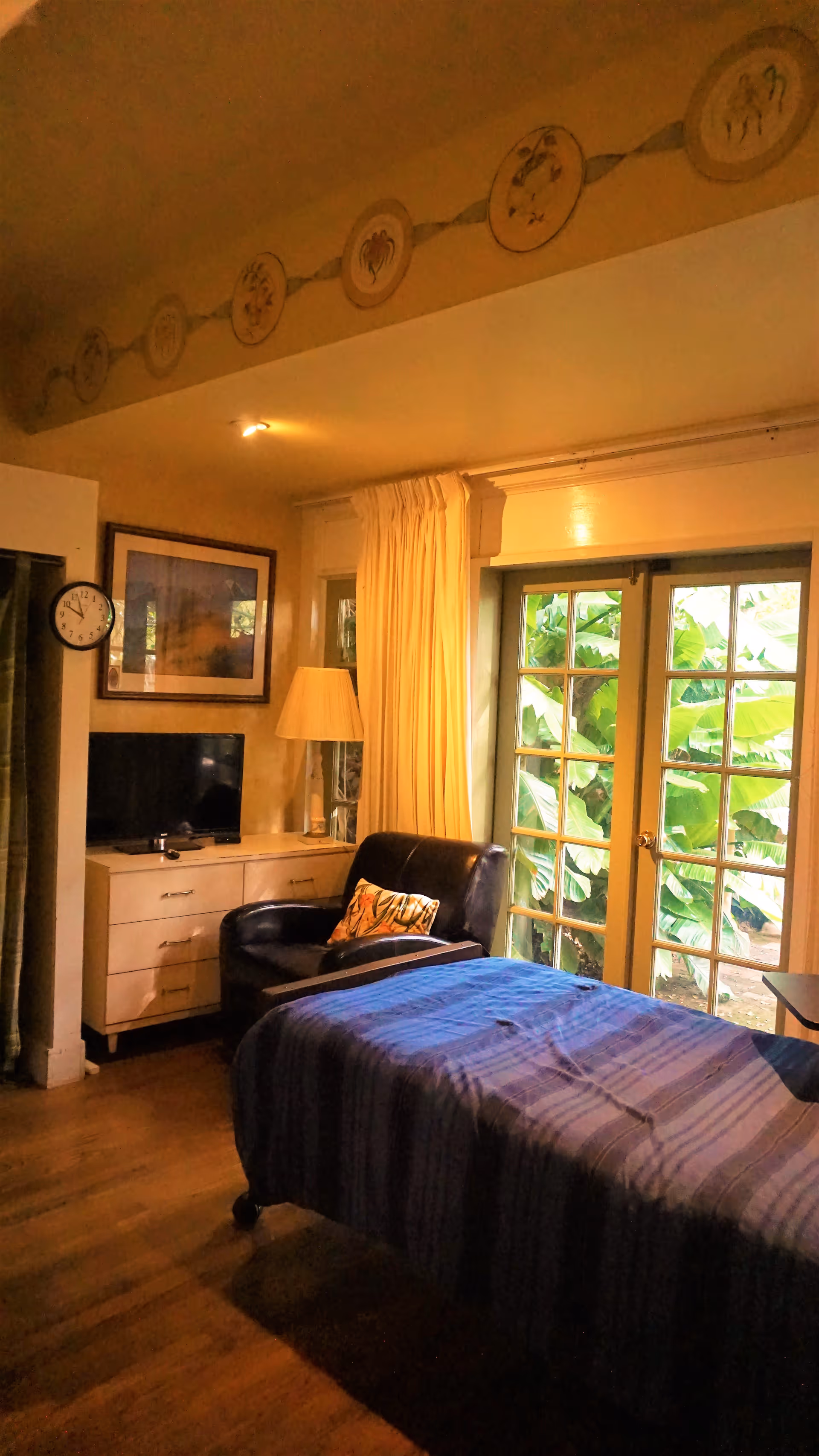 Private bedroom with a bed, dresser and TV, an armchair and lamp beside French doors opening to garden greenery.