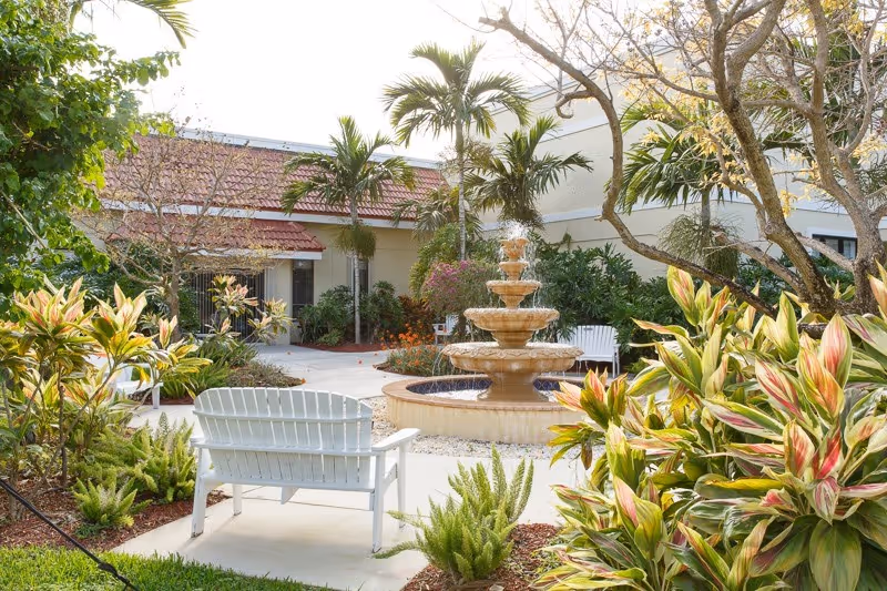 A peaceful outdoor courtyard with a three-tiered water fountain in the center, surrounded by lush tropical plants and trees. White benches are placed along the paved walkway, and a building with a red-tiled roof is visible in the background.