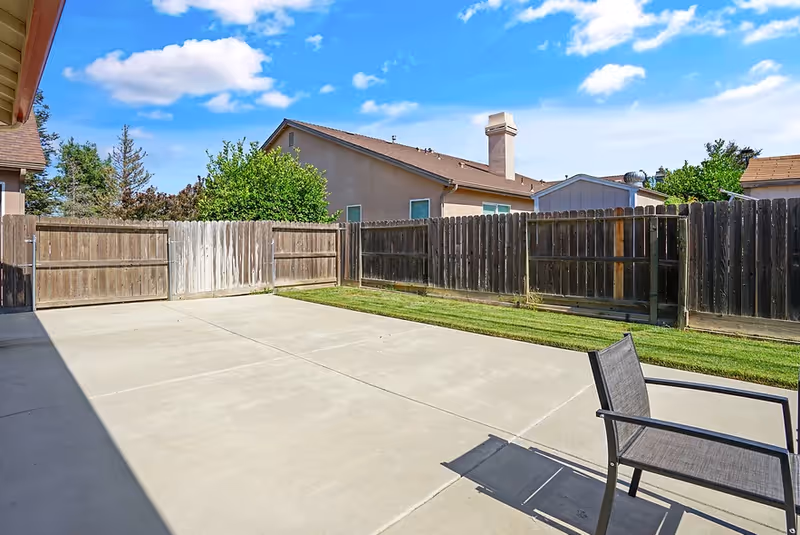 A fenced outdoor patio area with a concrete floor and a single chair. The patio is adjacent to a grassy strip and surrounded by wooden fences. Neighboring houses and a partly cloudy blue sky are visible in the background.