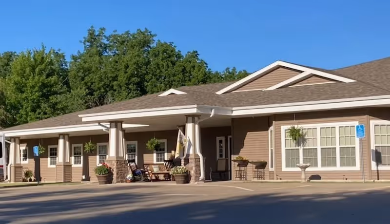 Front exterior of a single-story senior living facility with a covered entrance, columns, rocking chairs, potted plants, and handicap parking signs.