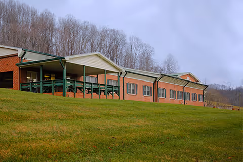 Exterior view of a single-story brick building with a covered porch area, surrounded by a grassy lawn and leafless trees in the background under a cloudy sky.
