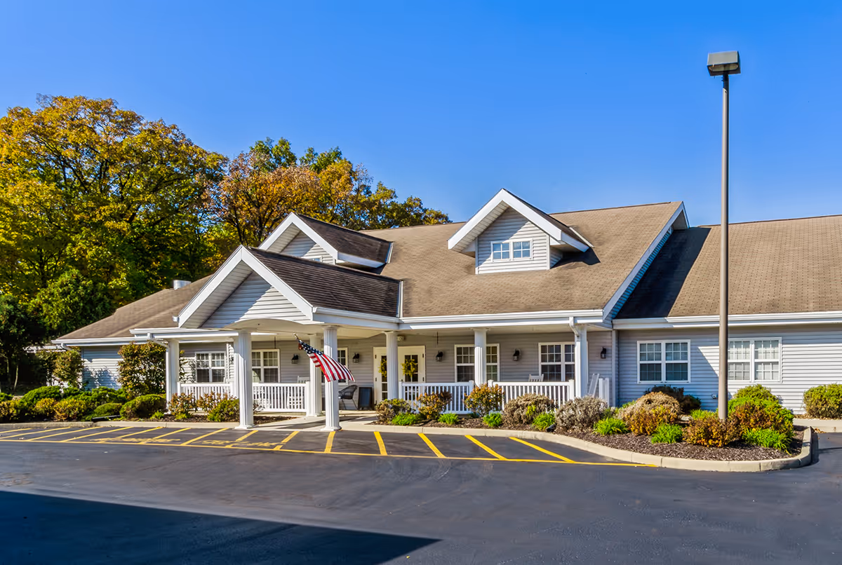Single-story gray senior living building with a covered porch entrance, American flag, landscaped beds, and a parking lot under a clear blue sky.
