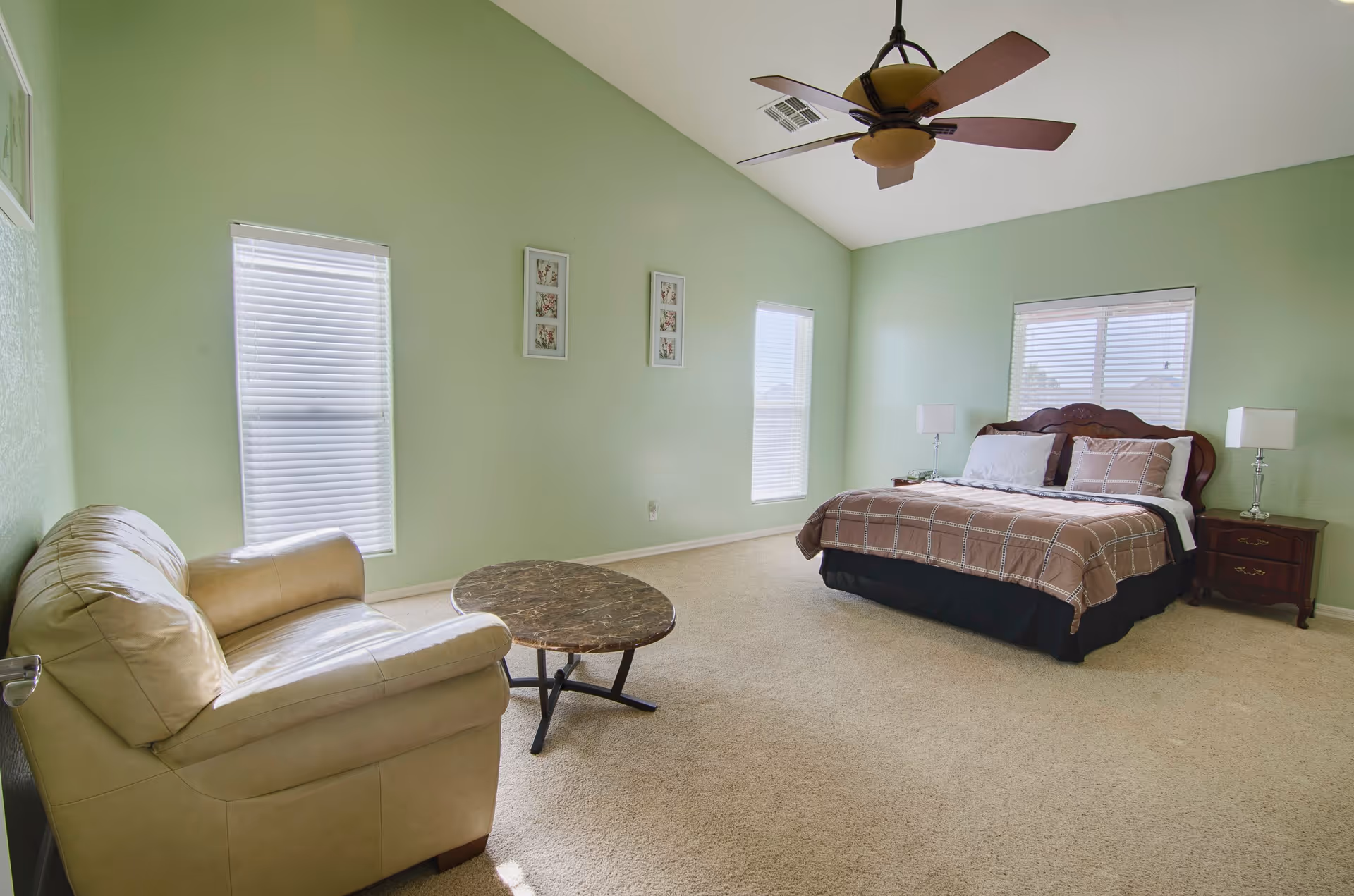 A spacious bedroom with light green walls and beige carpet. The room features a large bed with a brown checkered comforter, two wooden nightstands with lamps, a beige leather armchair, and a round marble-top coffee table. There are three windows with white blinds and three framed floral pictures on the wall. A ceiling fan with wooden blades is mounted on the white ceiling.