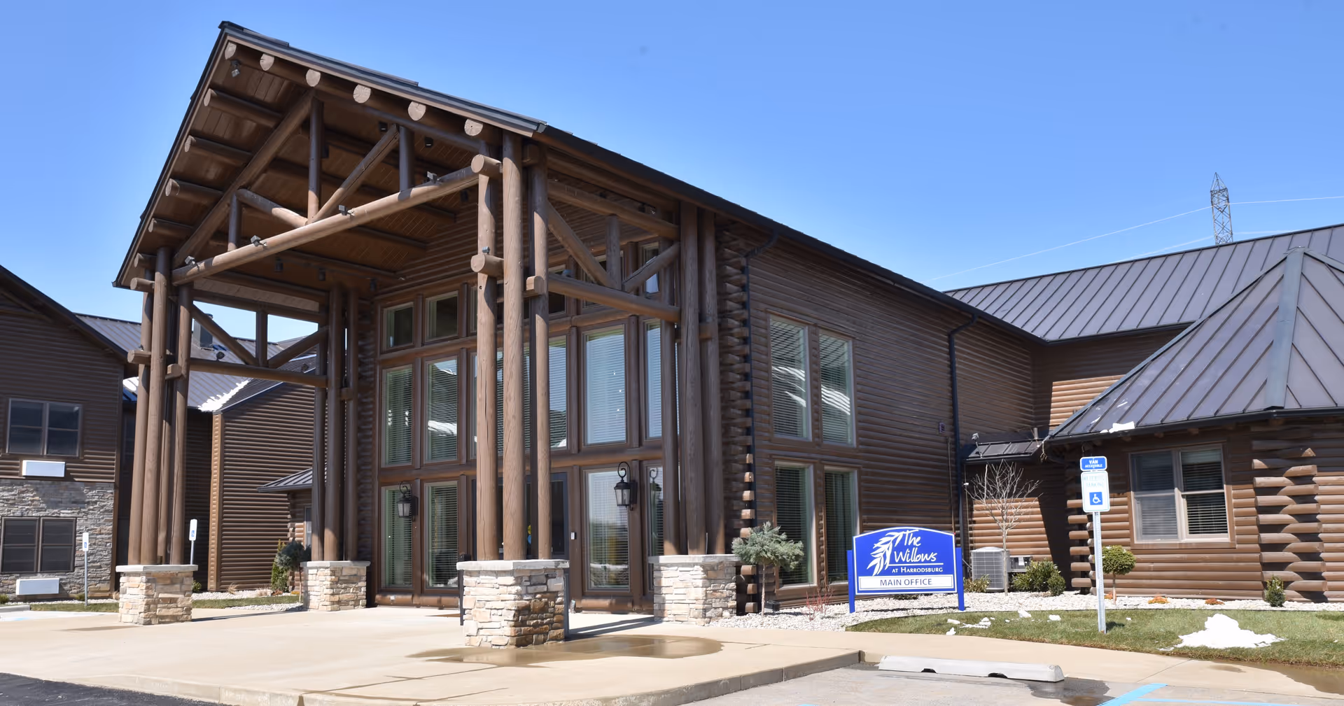 Entrance of a log-style senior living building with large timber columns, stone accents, and a sign for The Willows at Harrodsburg.