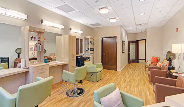 Interior view of a salon area in a senior living facility with light green salon chairs, wooden cabinets, mirrors, and hair care products. The room has wood flooring, beige walls, and a seating area with brown and green chairs along the right side.