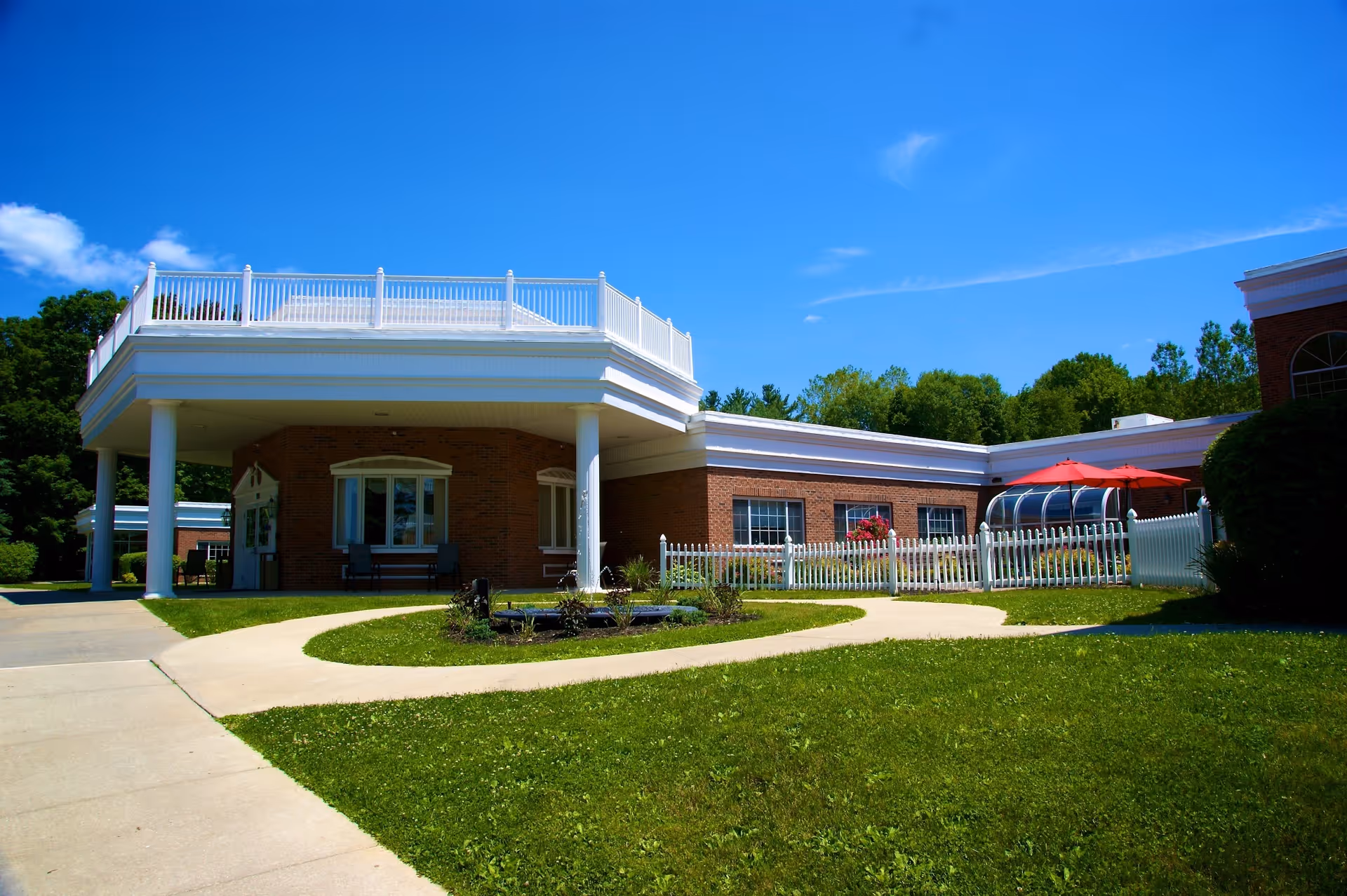 Brick single-story building with a covered columned entrance, rooftop white railing, white picket fence, lawn, and red patio umbrellas.