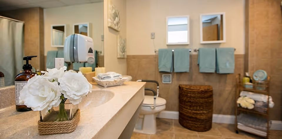 A clean and well-organized bathroom with a beige countertop holding a vase with white flowers and a soap dispenser. The bathroom features a toilet with a grab bar, a wicker laundry basket, two teal towels hanging on the wall, a small shelving unit with towels and toiletries, and two square mirrors above the towels.