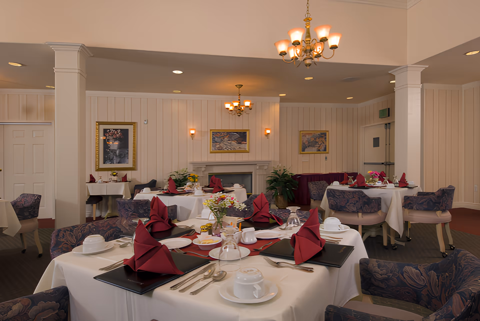 A dining room with several round tables covered with white tablecloths, each set with burgundy folded napkins, plates, cups, and silverware. The room has patterned upholstered chairs, a fireplace with framed artwork above it, wall sconces, and chandeliers providing warm lighting. There are plants and additional framed pictures on the paneled walls.