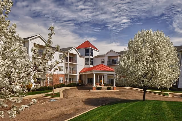 Exterior view of Sprenger Health Care Grande Village showing a multi-story building with white siding and red roofs. The entrance features a covered driveway with columns. The surrounding area includes blossoming trees and well-maintained landscaping under a partly cloudy sky.