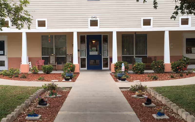 Front entrance of a building with a blue door, flanked by windows and white columns. There are chairs and benches on the porch area, with landscaped flower beds and small shrubs on either side of the walkway leading to the door.