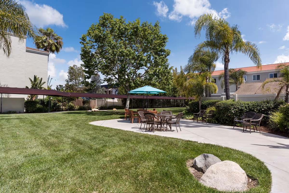 Outdoor patio area with several tables and chairs, including one table with a blue umbrella, surrounded by green grass, palm trees, and other plants. Residential buildings are visible in the background under a partly cloudy blue sky.