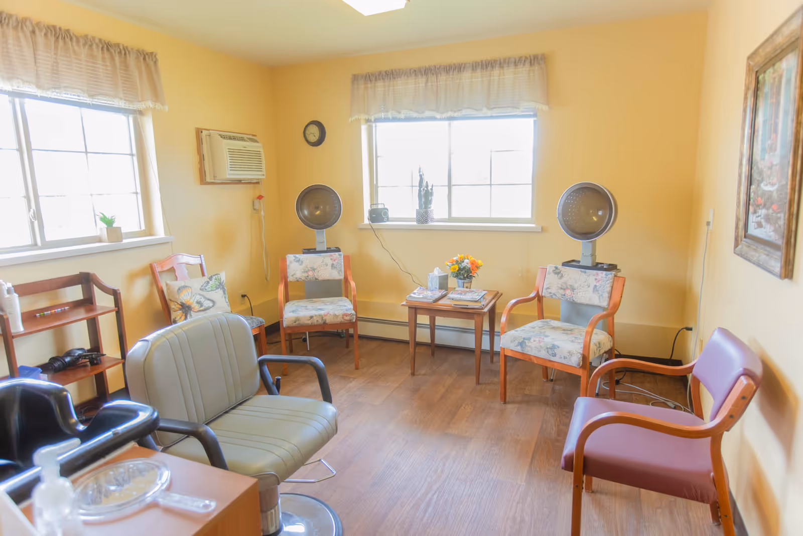 A bright room with yellow walls featuring two windows with sheer curtains, several chairs including salon chairs and floral upholstered chairs, two vintage hair dryers, a small wooden table with magazines and a flower vase, a wall clock, an air conditioning unit, and a framed painting on the wall.