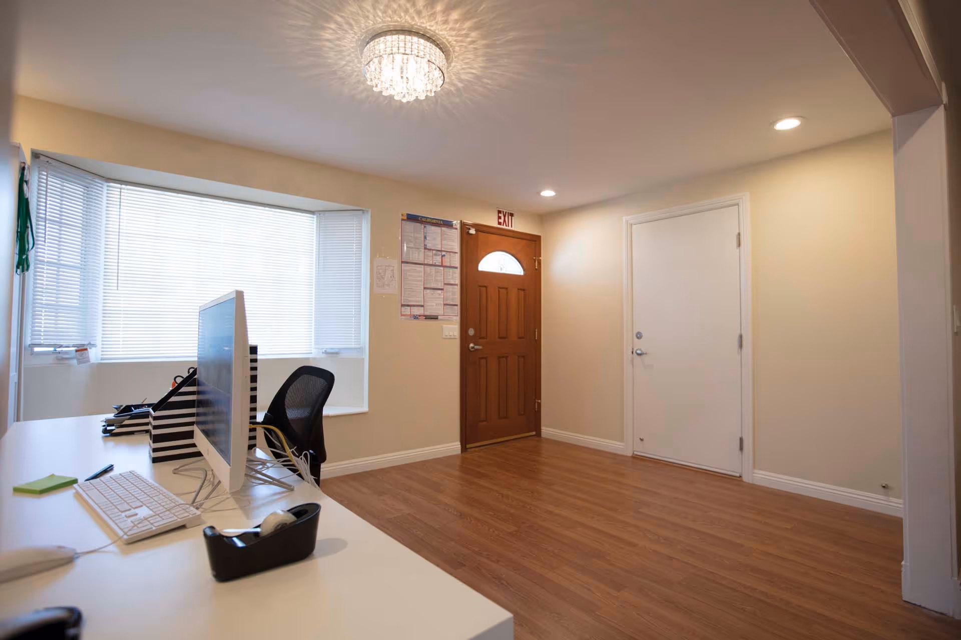 Interior view of an office or reception area with a wooden front door, a white door, a desk with a computer monitor, keyboard, and office supplies, and a large window with blinds letting in natural light. The floor is wooden, and the ceiling has a decorative light fixture.