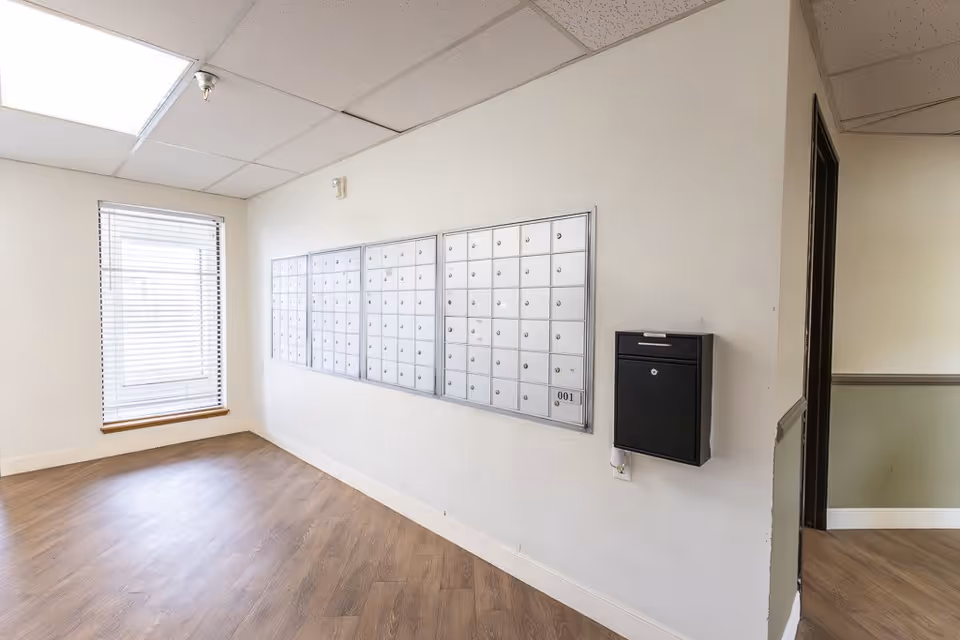 Interior view of a mailroom area in a senior living facility with multiple silver mailboxes mounted on a white wall and a black mailbox attached beside them. There is a window with blinds on the left side and wood-patterned flooring.