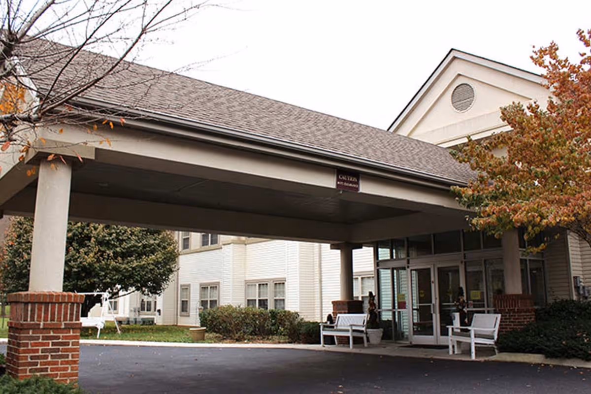 Entrance of Cedar Hills Senior Living facility showing a covered drop-off area with white benches on either side, brick pillars, and glass doors leading inside. Trees with autumn leaves and shrubs surround the building.