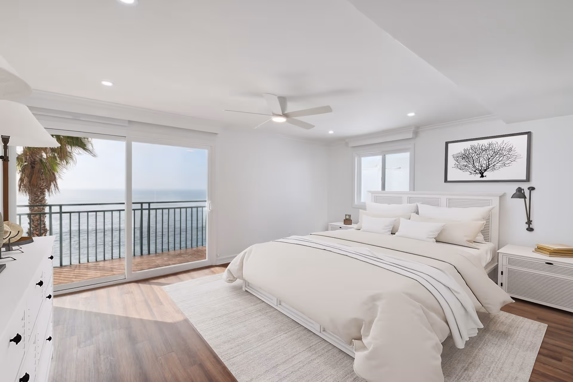 Bright and airy bedroom with a large bed dressed in white linens, two nightstands, a dresser, a ceiling fan, and a sliding glass door leading to a balcony overlooking the ocean with a palm tree visible outside.