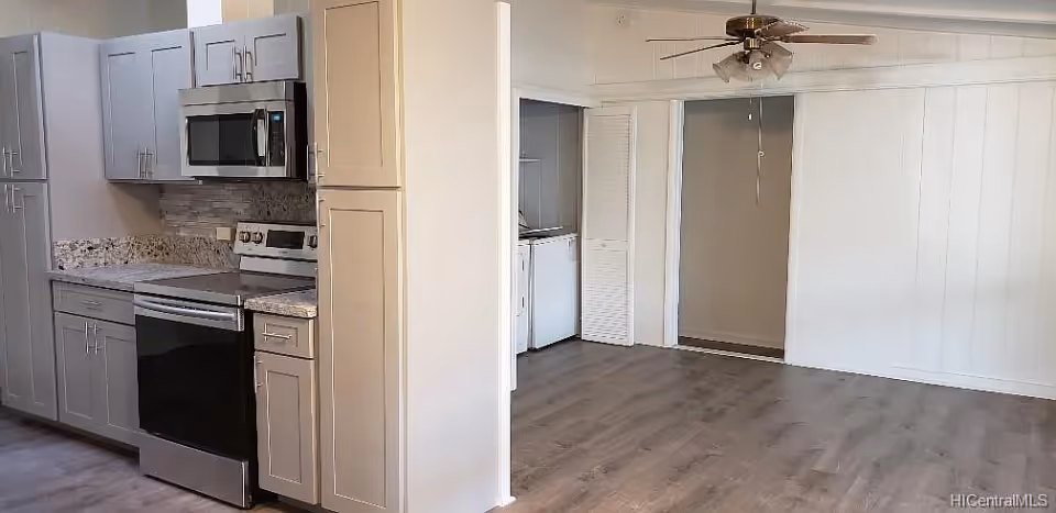 Interior view of a modern kitchen and adjacent empty living area with wood flooring, white cabinets, stainless steel appliances including a microwave and stove, and a ceiling fan in the living area.