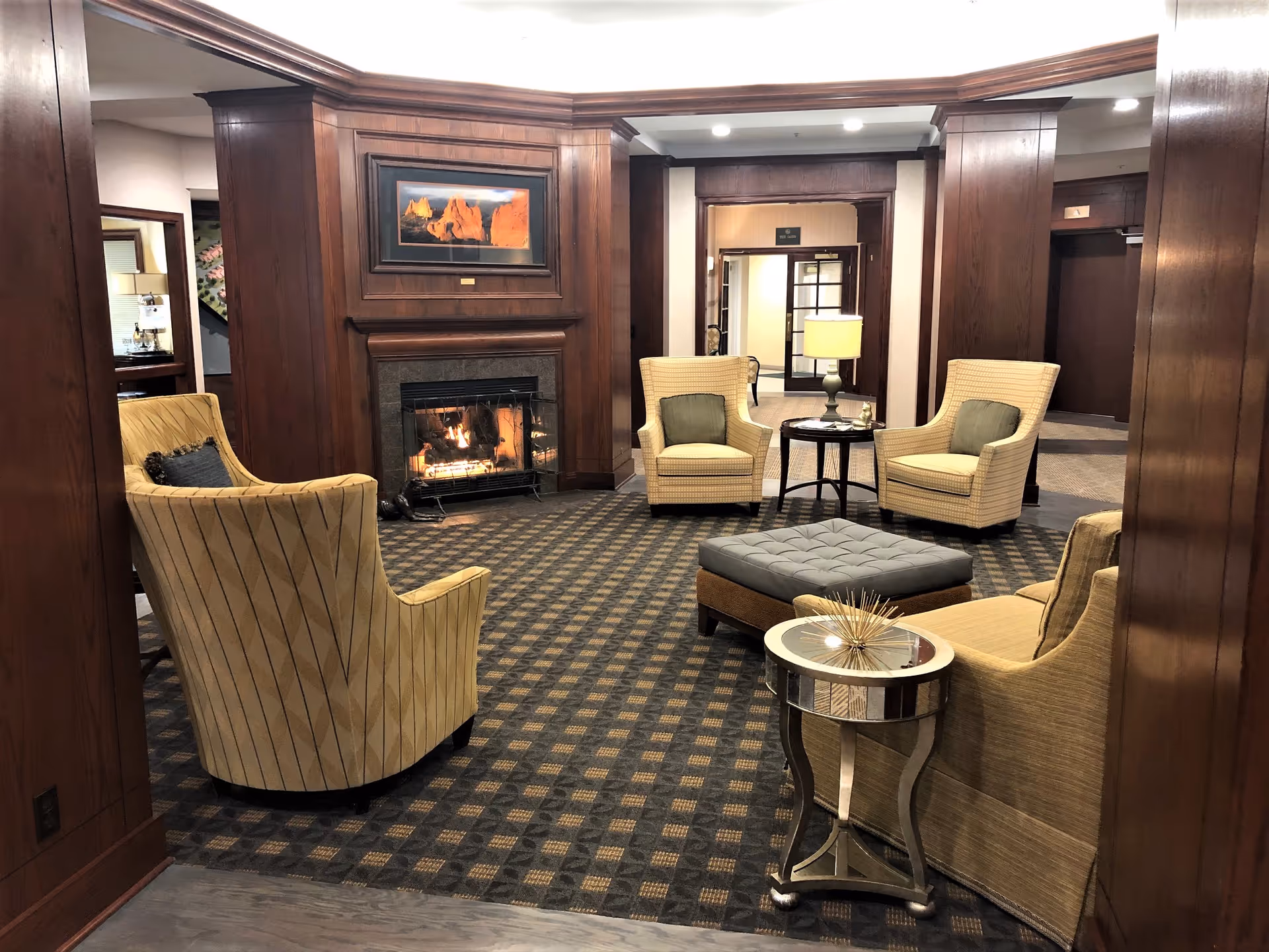 A cozy seating area in a senior living facility with four upholstered armchairs arranged around a square ottoman and a small round side table. The room features a lit fireplace set in a wooden paneled wall with a framed landscape painting above it. The carpet has a checkered pattern, and there is a lamp on a table in the background near a doorway.