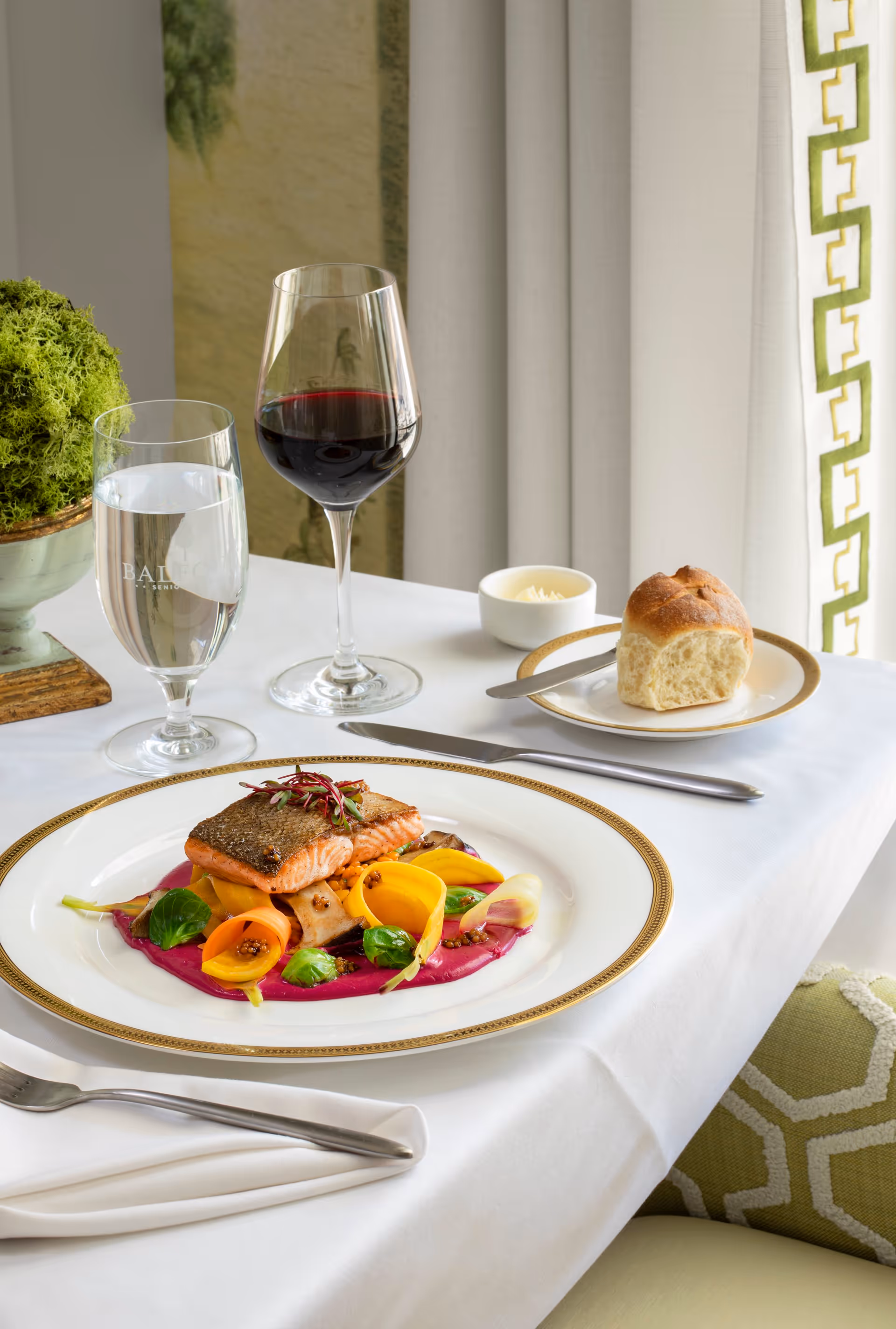 A fine dining table setting with a plated salmon dish garnished with colorful vegetables and sauce, a glass of red wine, a glass of water, a bread roll on a small plate with butter, and silverware on a white tablecloth near a window with curtains.