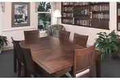 A room with a large wooden table surrounded by eight wooden chairs. In the background, there is a bookshelf filled with books, a floor lamp, a framed picture on the wall, and a window with a view of greenery outside. There are also two potted plants on either side of the table.