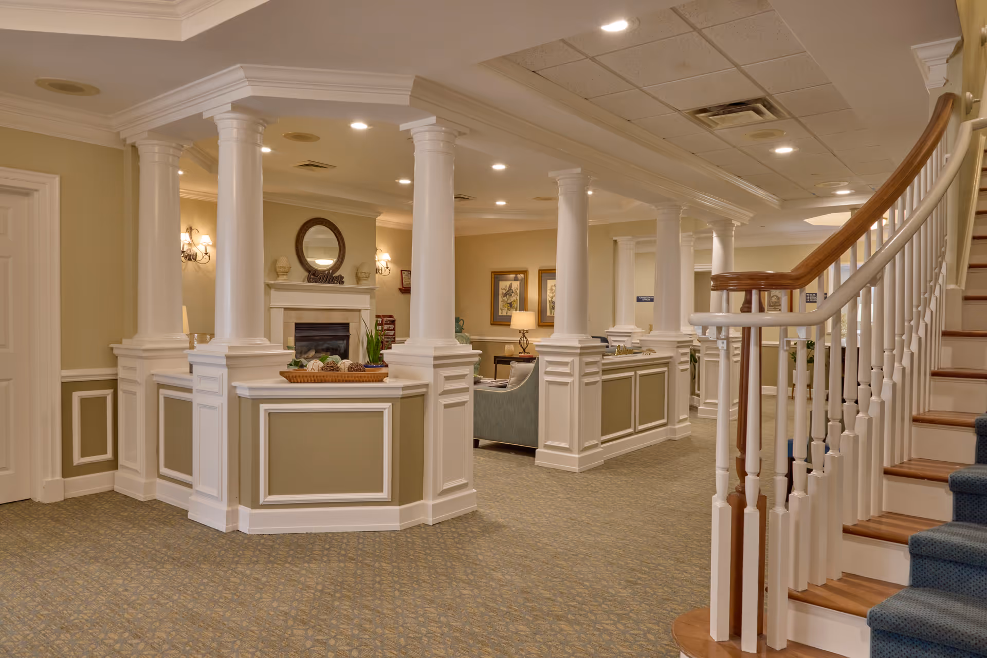 Elegant senior living facility lobby with white columns, a seating area by a fireplace, and a curved staircase.