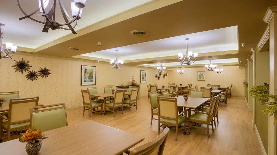 A spacious dining room with multiple wooden tables and green cushioned chairs arranged neatly. The room features warm yellow walls with framed bird artwork and decorative sunburst wall hangings. Several chandeliers with multiple lights hang from the ceiling, illuminating the room. The floor is wooden, and there are small flower arrangements on the tables.