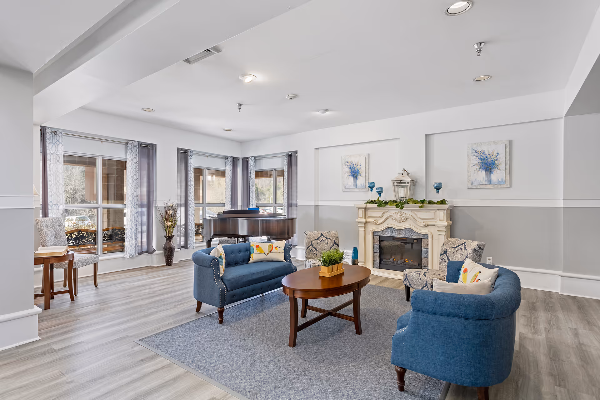 A bright and spacious living room area with light gray walls and wood flooring. The room features a blue upholstered sofa and two matching armchairs arranged around a wooden coffee table on a gray area rug. There is a decorative white fireplace with blue and white artwork above it, and a grand piano near large windows with sheer patterned curtains allowing natural light to fill the space.