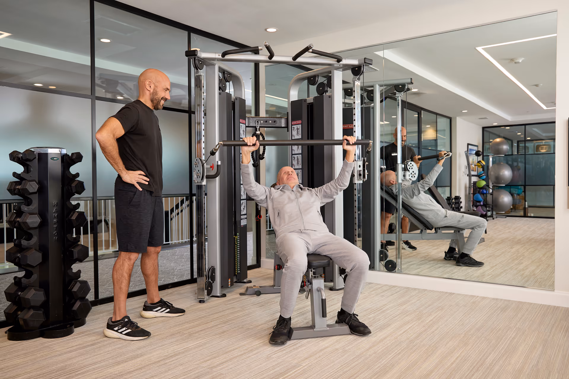 An elderly man in a gray tracksuit is using a weight machine for bench pressing in a fitness room, while a younger man in black athletic wear stands beside him, offering support. The room has a rack of dumbbells, large mirrors on the wall, and exercise balls in the background.