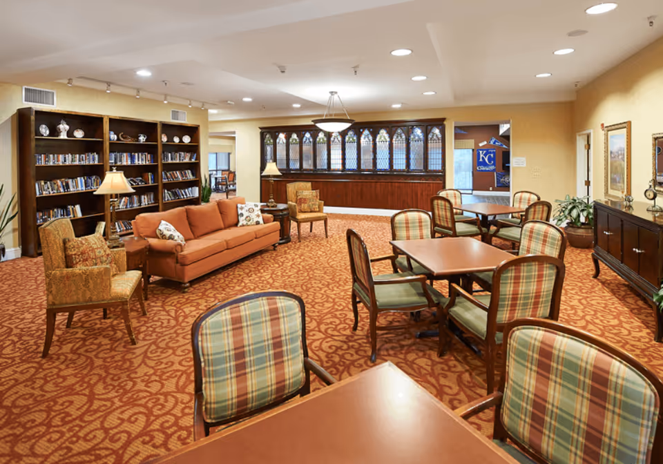 A spacious senior living common area with patterned carpet, several tables and chairs with plaid upholstery, a comfortable orange sofa, armchairs, bookshelves filled with books, and warm lighting from lamps and ceiling lights.