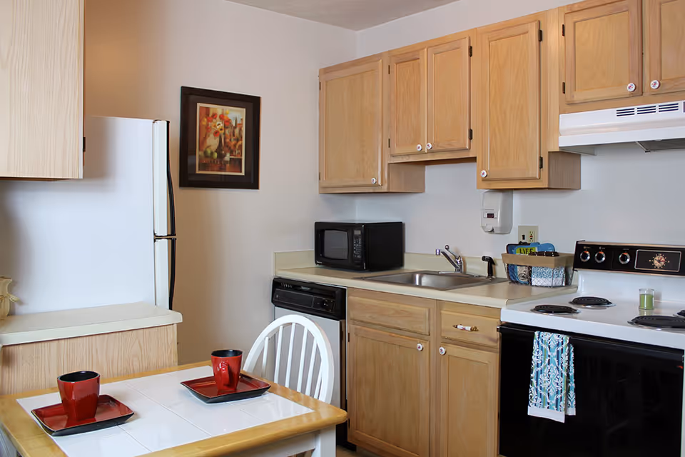 A small kitchen area with light wood cabinets, a white refrigerator, a black microwave, a white stove with a patterned towel hanging on the handle, and a sink. A small dining table with two red cups and matching square plates is in the foreground. A framed floral painting hangs on the wall.