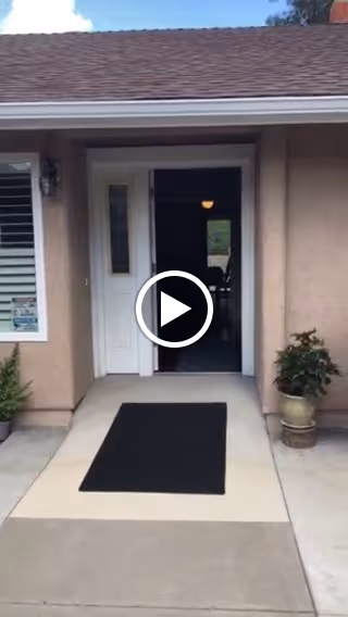 Front entrance of Mount Carmel Assisted Living with an open white door, black doormat, potted plants, and a short walkway.