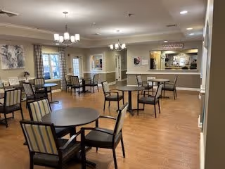 A spacious dining room with several round tables and chairs arranged neatly. The room has wooden flooring, beige walls, and large windows with curtains allowing natural light. There are two chandeliers hanging from the ceiling and a serving counter with a pass-through window in the background.