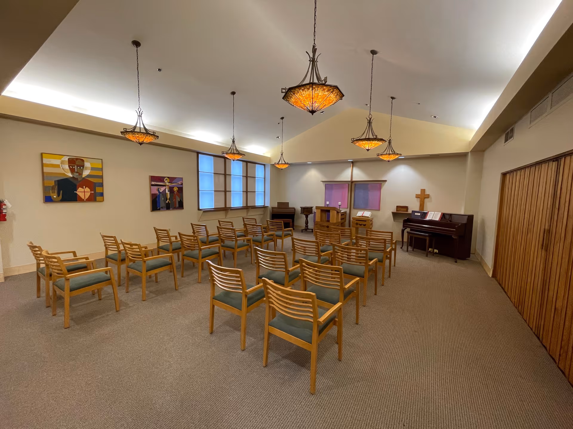Interior chapel with rows of wooden chairs facing a pulpit, piano, cross, and hanging pendant lights.
