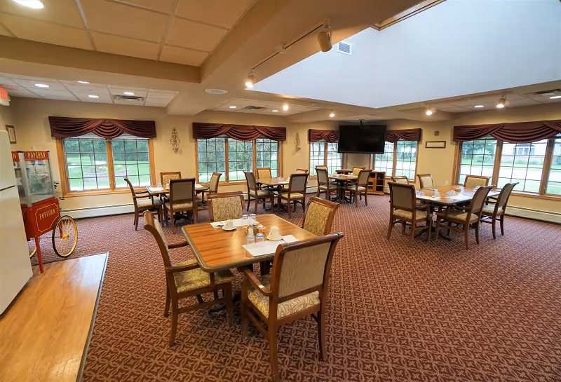 A spacious dining room with multiple wooden tables and cushioned chairs arranged neatly. Each table is set with placemats, cups, and condiments. Large windows with maroon valances allow natural light to fill the room. A popcorn machine is visible on the left side near the entrance, and a flat-screen TV is mounted on the far wall. The room has a patterned carpet and a skylight in the ceiling.