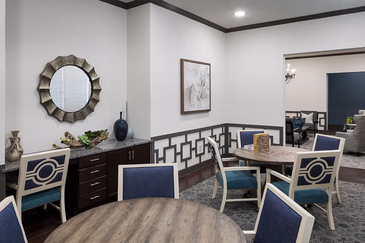 Decorated common dining/activity room with round wooden tables, blue-upholstered chairs, a decorative mirror and vases on a sideboard.
