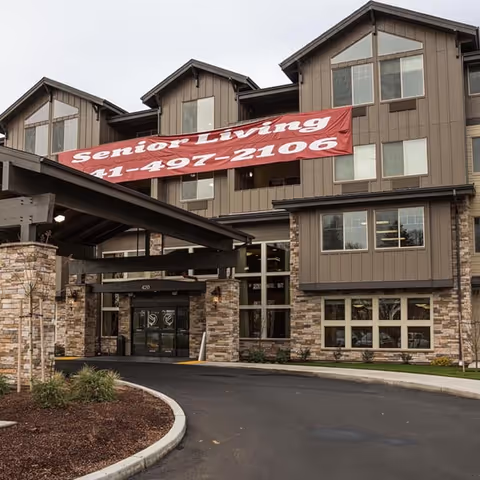 Exterior view of a senior living facility building with stone and wood paneling. A large red banner hangs above the entrance with the text 'Senior Living 41-497-2106'. The entrance has a covered driveway and landscaped areas with small plants and mulch.