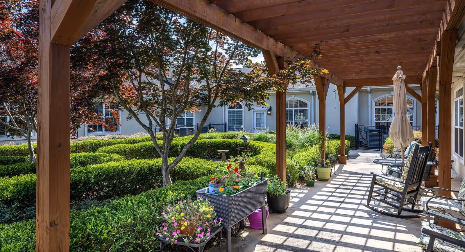 A shaded outdoor patio area with wooden pergola overhead, several rocking chairs, potted plants, and a flower bed. In the background, there is a well-maintained garden with trimmed hedges and trees, and the exterior of a building with windows and doors.