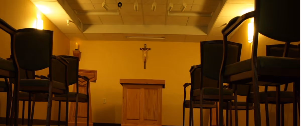 Interior of a small chapel or prayer room with wooden chairs arranged in rows facing a wooden podium and a crucifix mounted on the wall. The room is softly lit with wall sconces and ceiling lights.