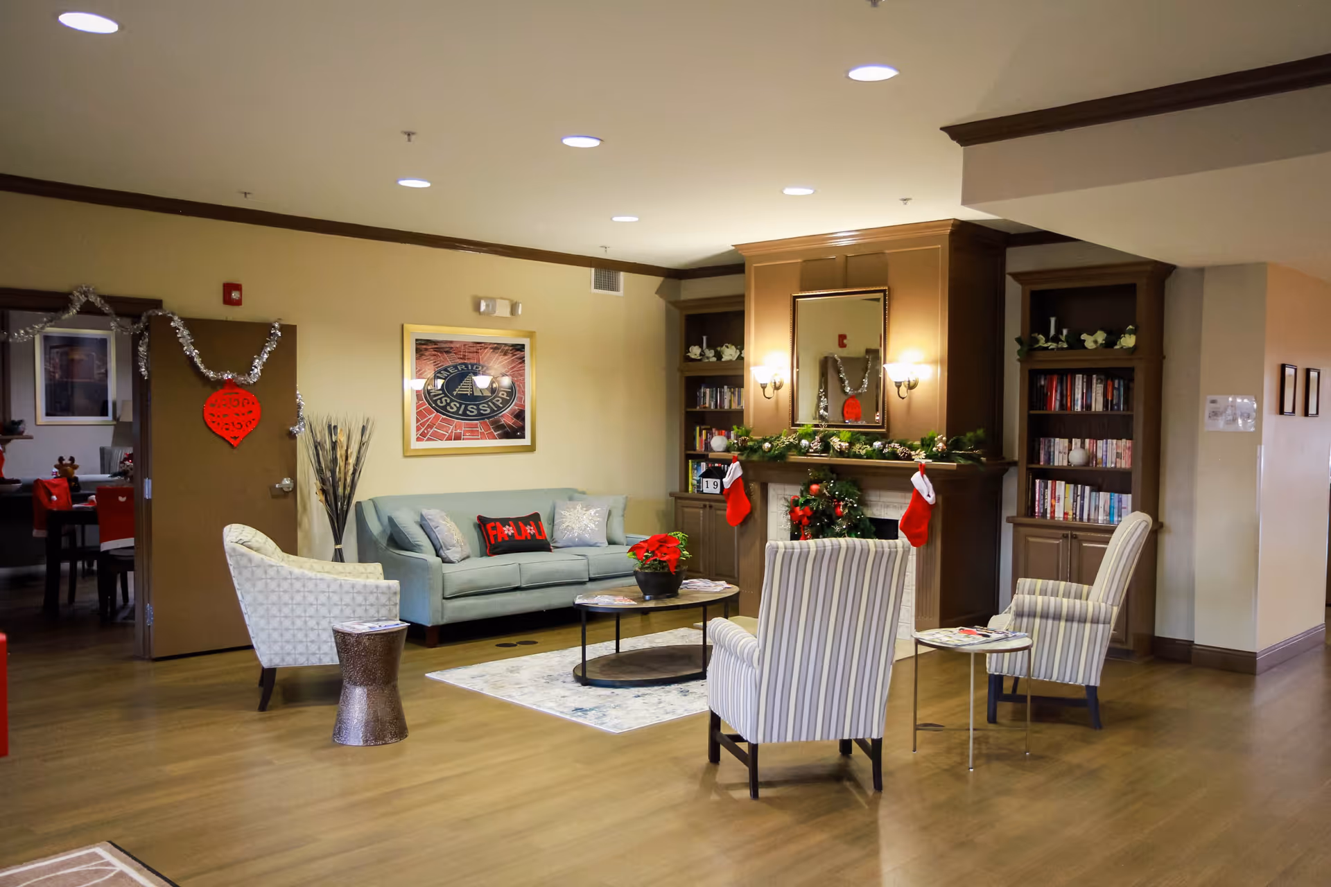 A cozy living room area decorated for Christmas with a light blue sofa, two striped armchairs, a round coffee table with a poinsettia plant, and a fireplace adorned with stockings and garlands. Bookshelves flank the fireplace, and a framed picture hangs on the wall. The room has wooden flooring and warm lighting.