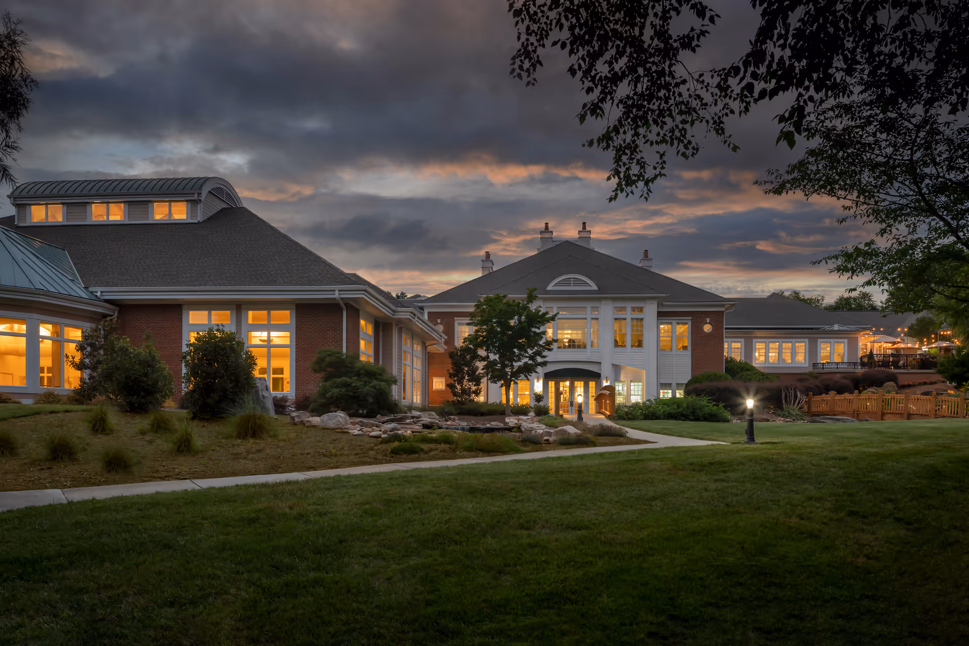 Exterior view of The Cypress of Charlotte senior living facility at dusk, showing a large building with many lit windows, a well-maintained lawn, trees, and a pathway leading to the entrance under a cloudy sky.