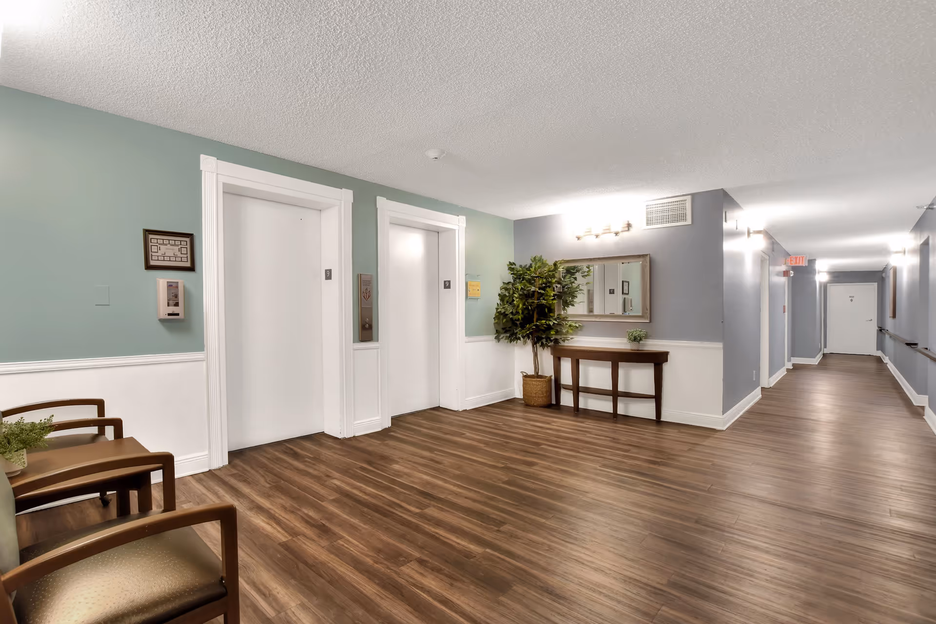 Interior hallway of a senior living facility with two white elevator doors marked with the number 9, wood flooring, light blue and white walls, a wooden table with a potted plant, a large mirror above the table, and a green leafy plant in a basket. There are chairs with armrests on the left side and an exit sign visible down the corridor.