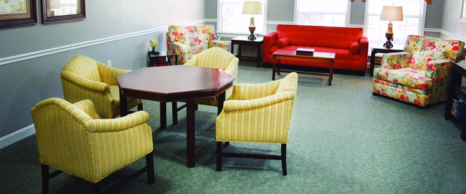 A senior living common room with a red sofa, floral armchairs, and four yellow patterned chairs around a wooden octagonal table.