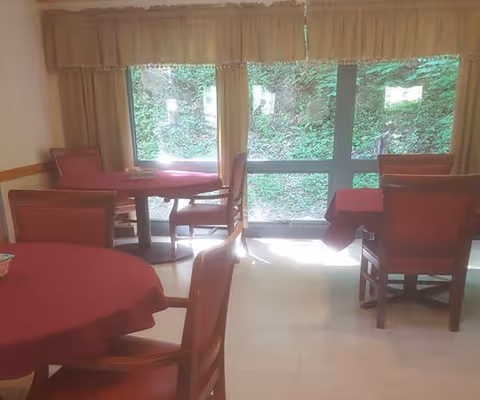 Interior view of a dining room with round tables covered in red tablecloths and wooden chairs. Large windows with beige curtains provide a view of green foliage outside.