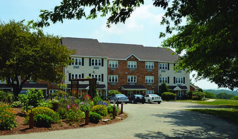 Exterior view of a multi-story senior living facility building with a brick and siding facade, surrounded by trees and landscaped garden beds. Several cars are parked in front of the building under a clear sky.