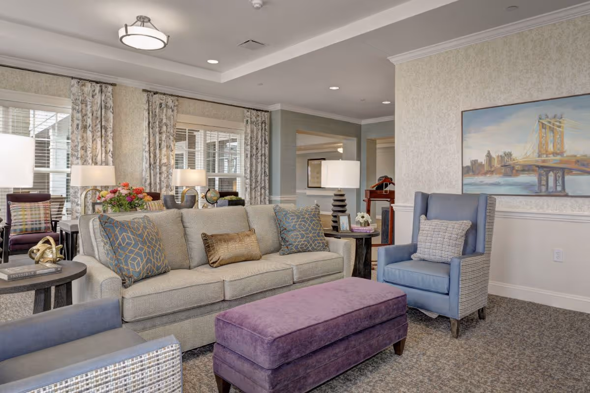 Bright, well-appointed lounge with a sofa, armchairs, a purple ottoman, table lamps, and wall art.