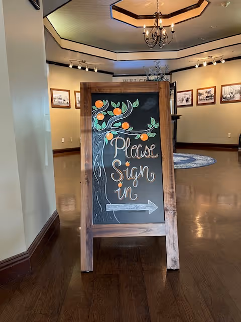A wooden-framed chalkboard sign in a lobby reads "Please Sign In" with painted oranges and an arrow, standing on a polished wood floor.